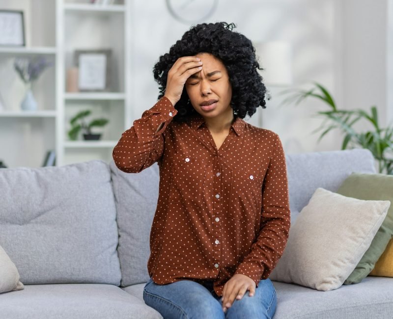 Worried young woman sitting on couch at home feeling stress, anxiety or headache